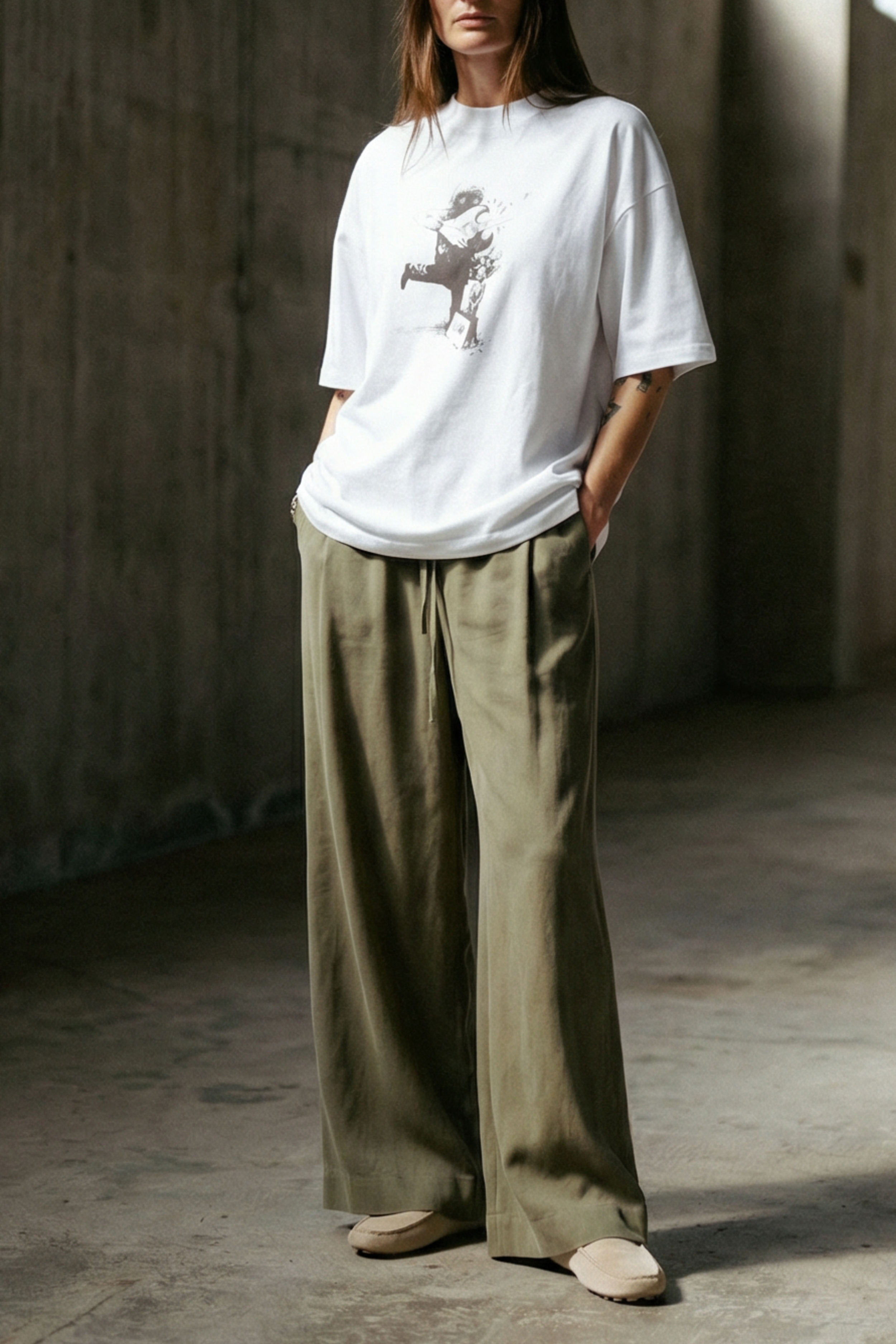 Person wearing a white t-shirt and beige pants standing in a sunlit concrete room.