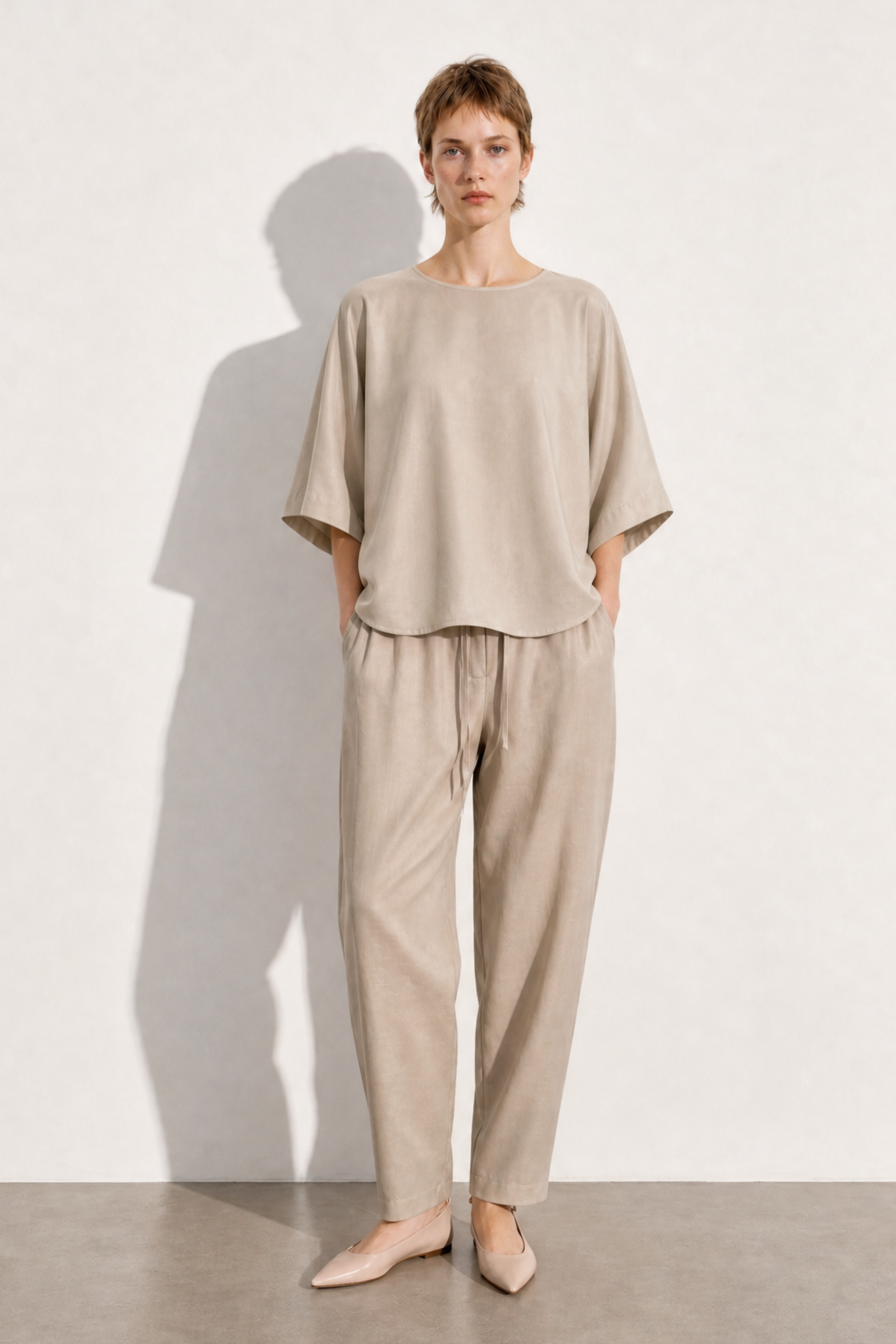 A model stands against a textured off-white wall wearing a matching beige linen two-piece set consisting of a short-sleeve top and relaxed-fit trousers, paired with beige pointed-toe flats.
