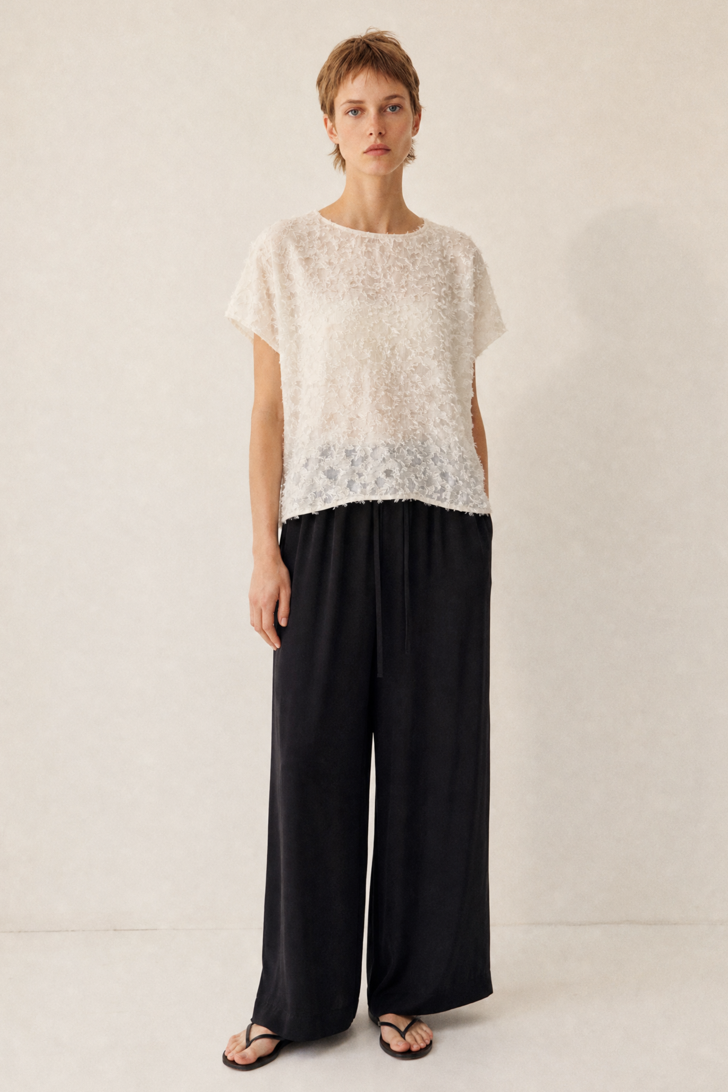 A model stands against a light beige textured wall wearing a cream-colored lace short-sleeve top and black wide-leg trousers.