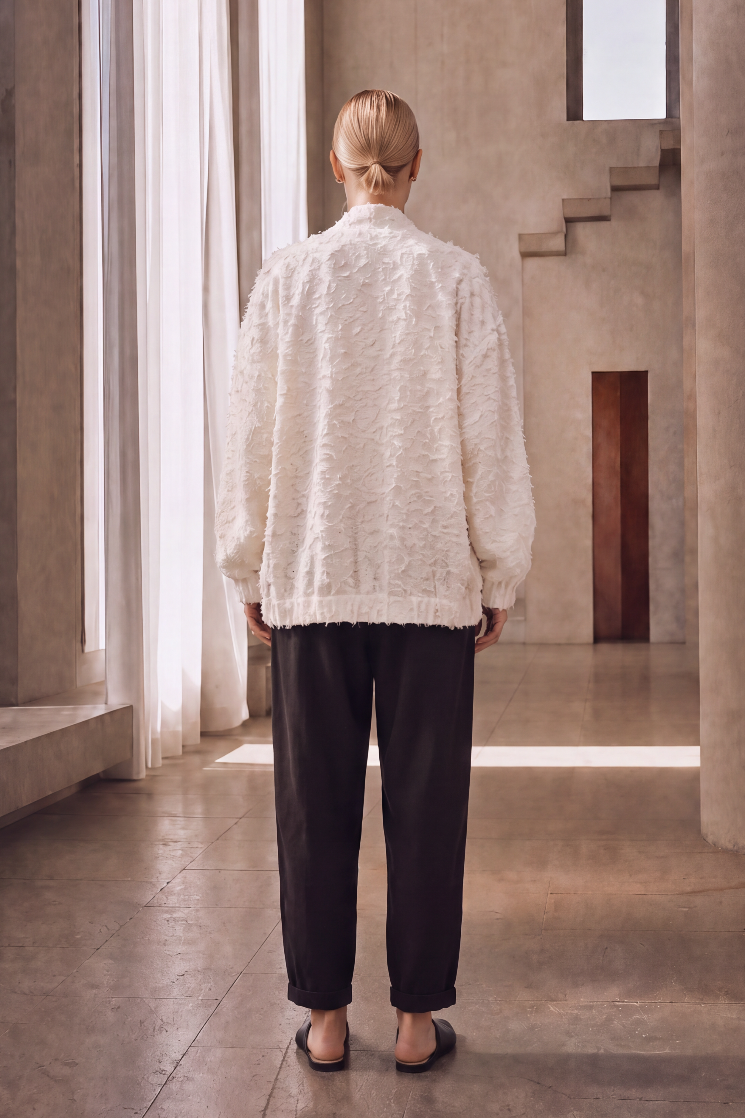 A woman viewed from behind wearing a textured cream-colored long-sleeve top and dark charcoal pants, standing in a minimalist concrete hallway with a staircase in the background.