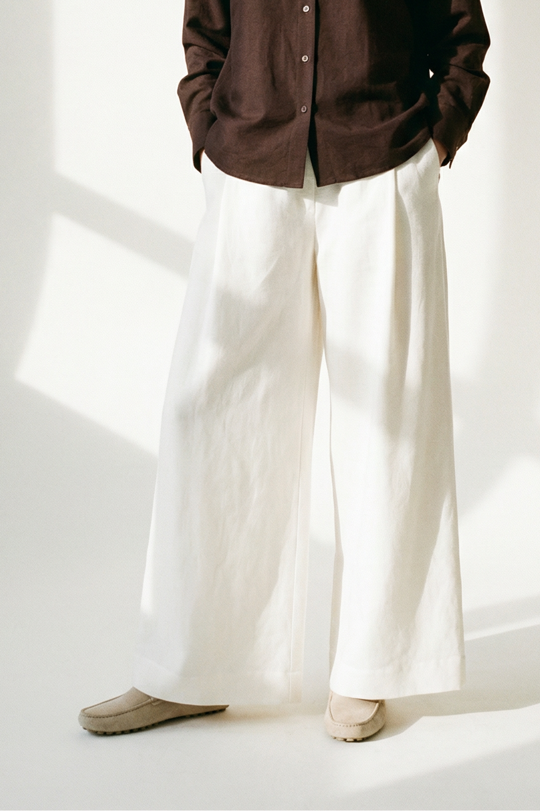 Woman wearing a brown shirt and white pants standing in a well-lit room with sunlight casting shadows on the wall.