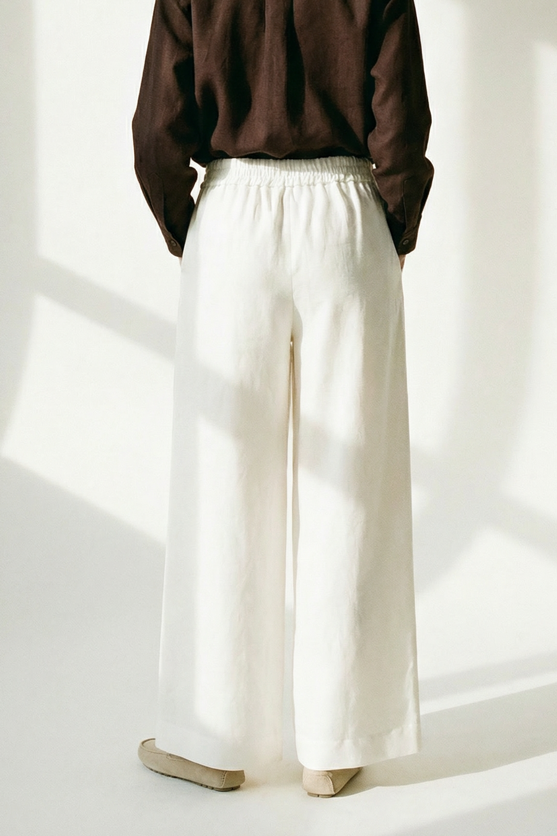 Person wearing a brown shirt and white pants standing in a well-lit room with sunlight casting shadows on the wall.