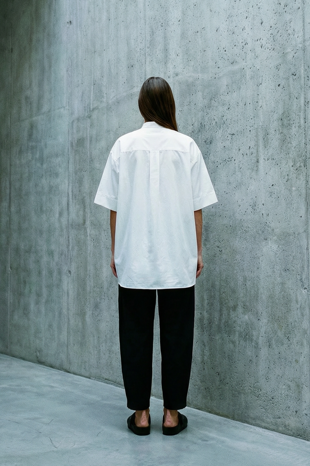 Person wearing a white shirt and black pants standing in a concrete hallway.