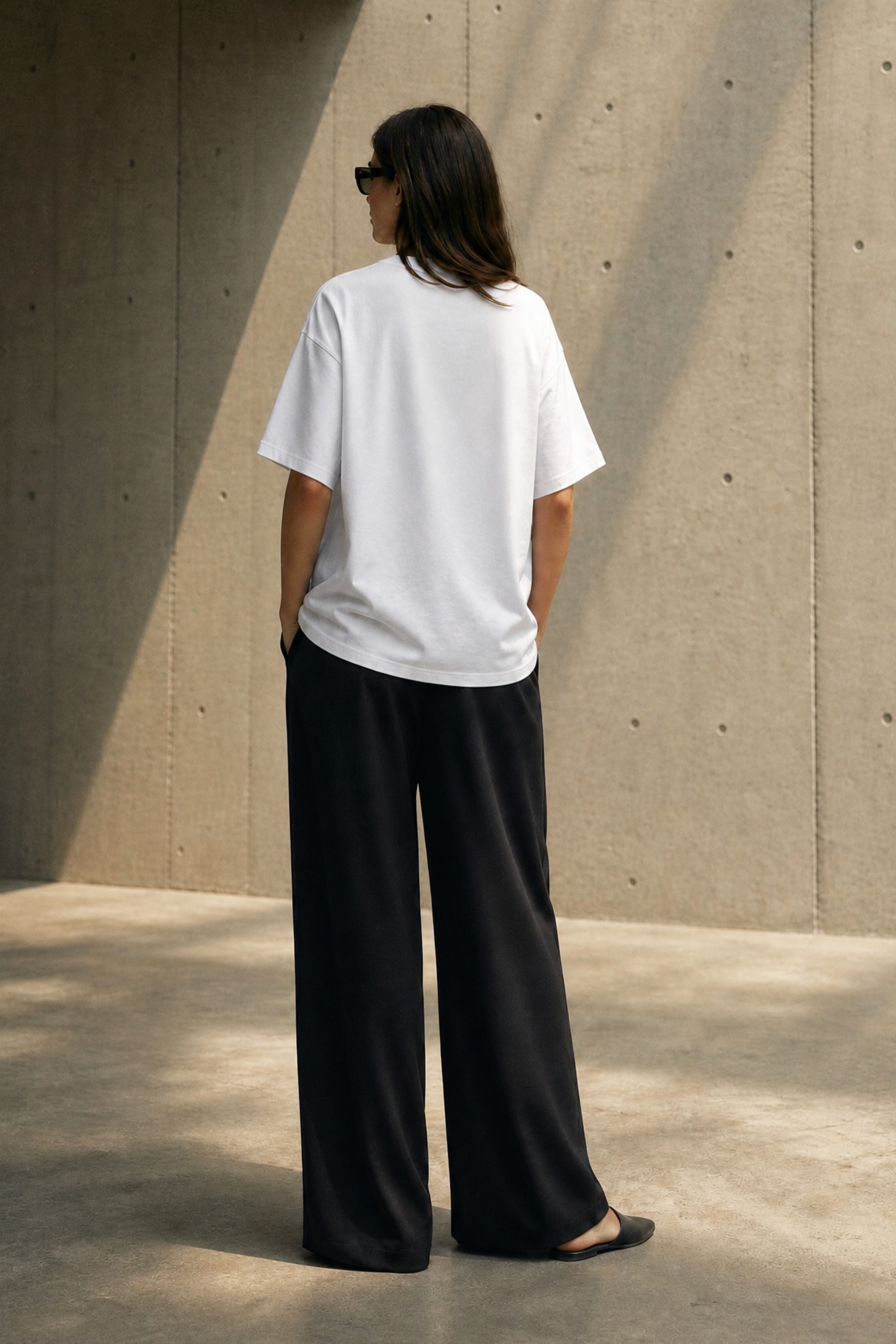Person wearing a white t-shirt and black pants standing against a concrete wall.
