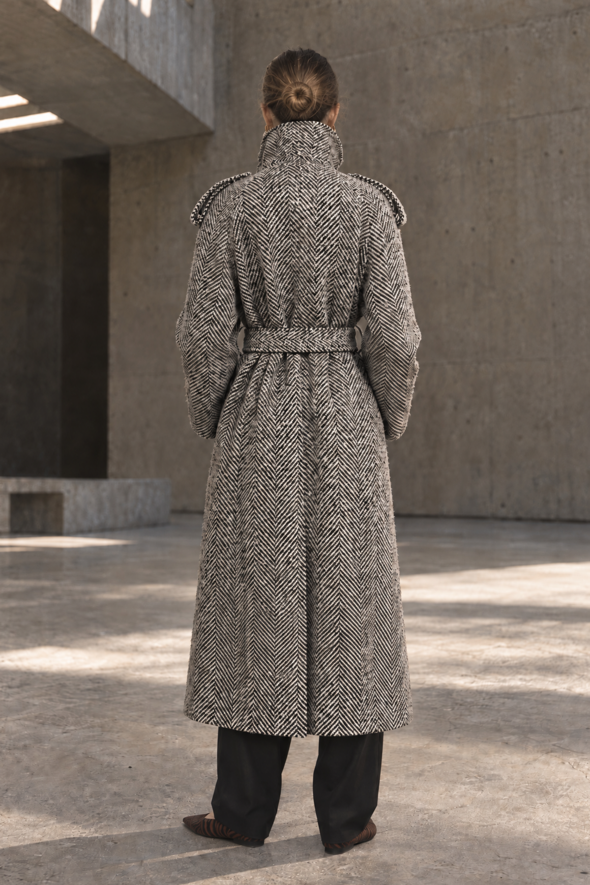 A person viewed from behind wearing a long herringbone-patterned coat in a concrete architectural setting.