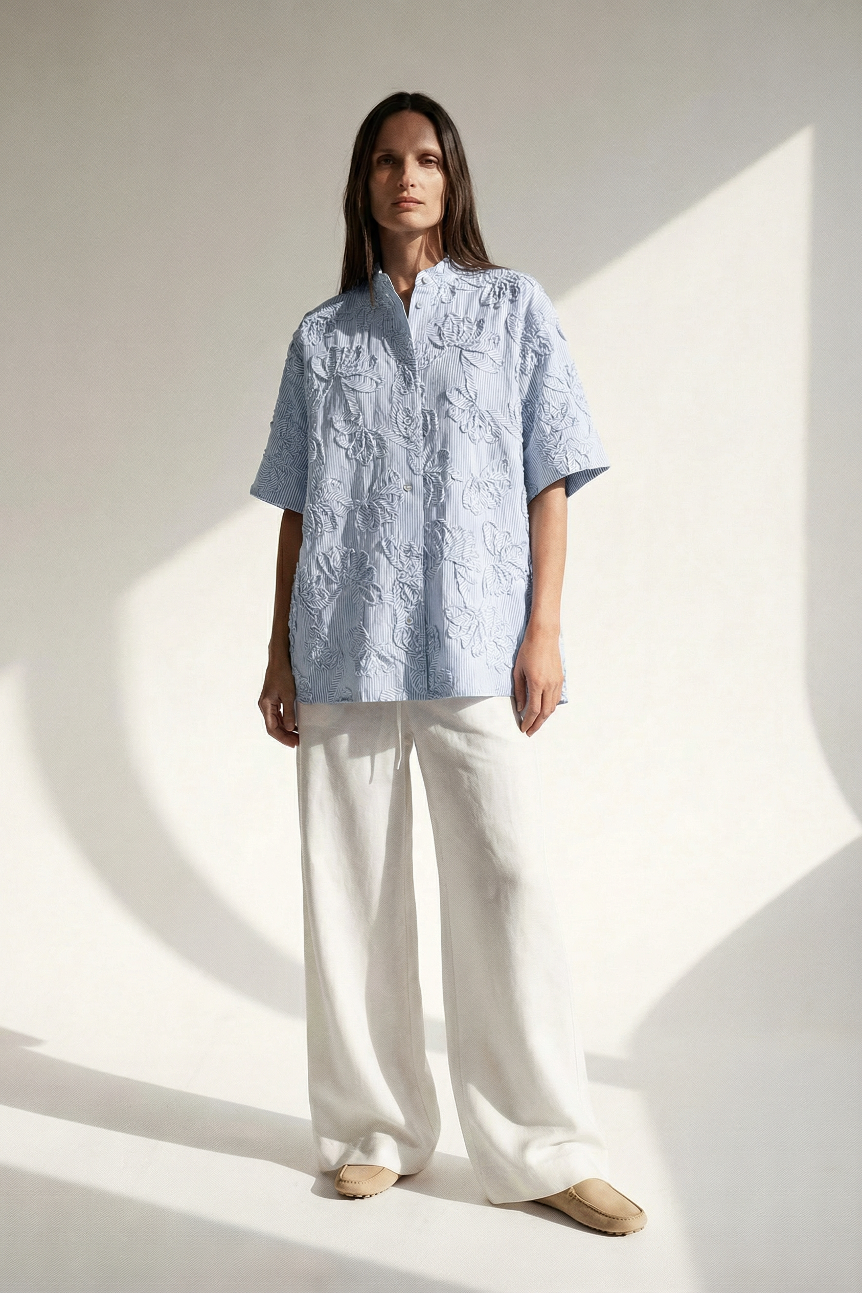 Woman wearing a light blue patterned shirt and white pants against a white background